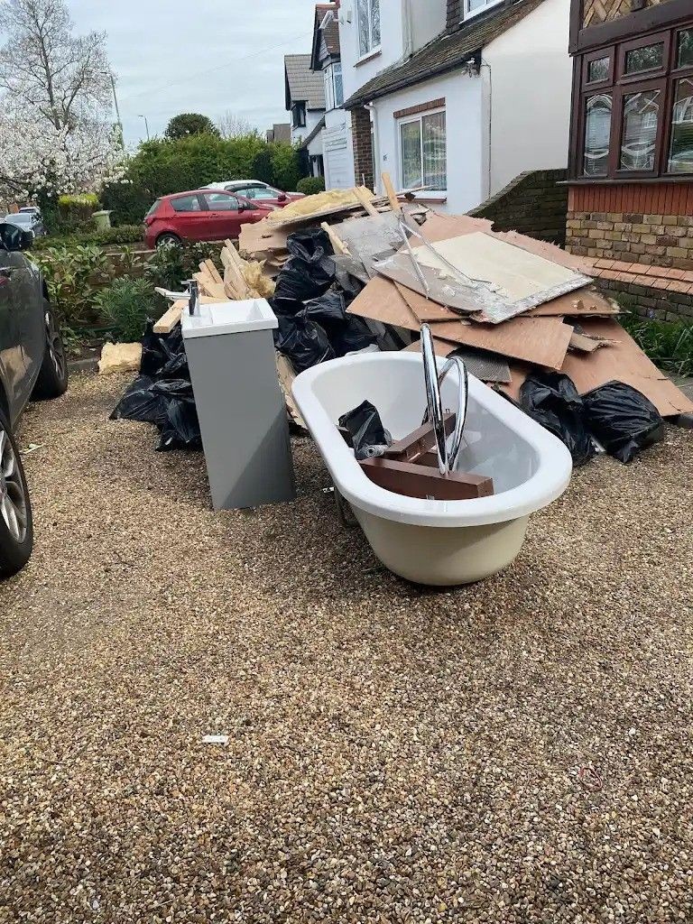 Pile of construction debris and an old bathtub on a gravel driveway in front of a residential house.