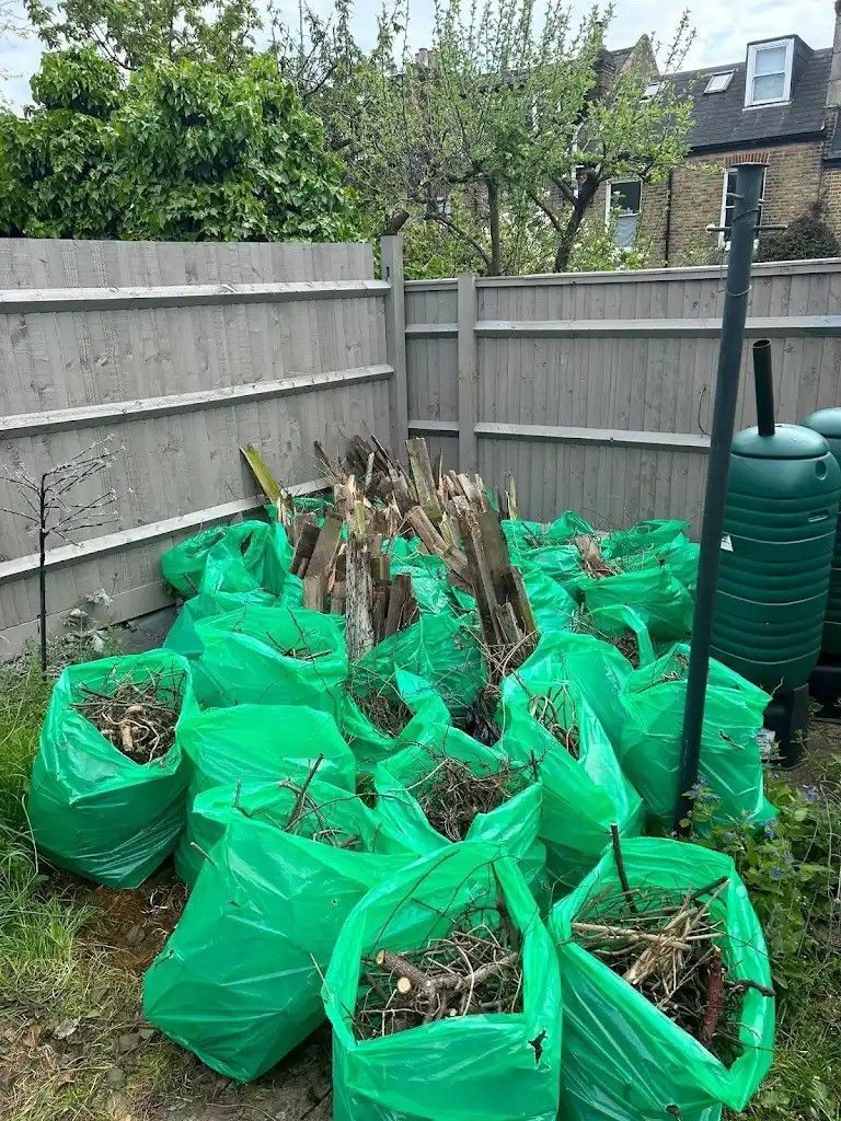 Green bags filled with garden waste and twigs against a wooden fence in a backyard.